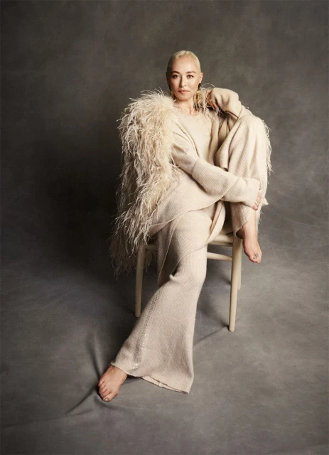 An adult woman with a shaved head poses seated on a chair in a neutral-colored sweater and trousers, featuring a feathered garment on her shoulders.